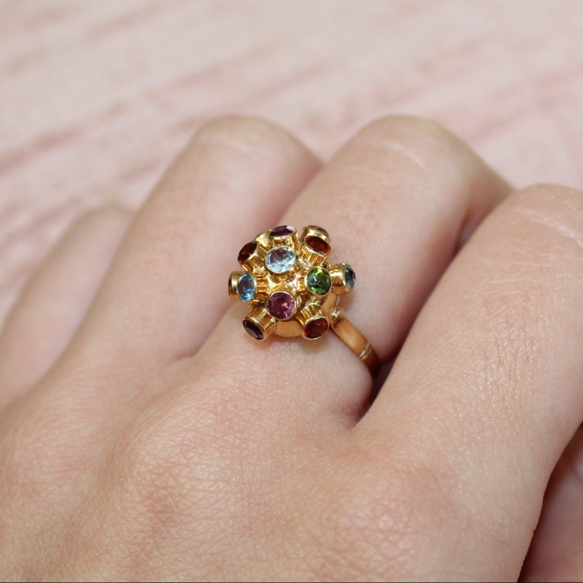 Gold ring with multicolored gemstones on a finger against a pink background