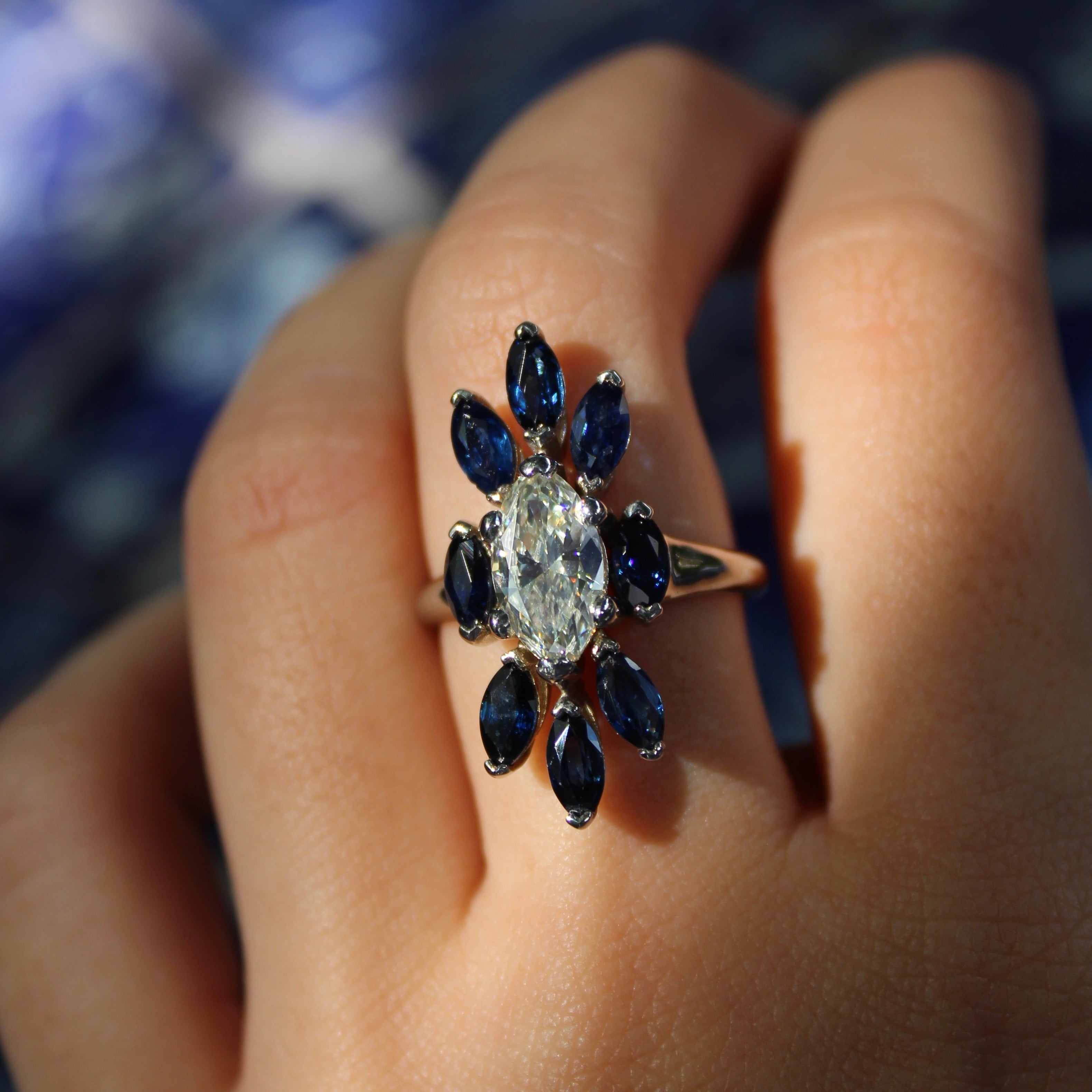 Ring with a central diamond and blue gemstones on a hand against a blurred background