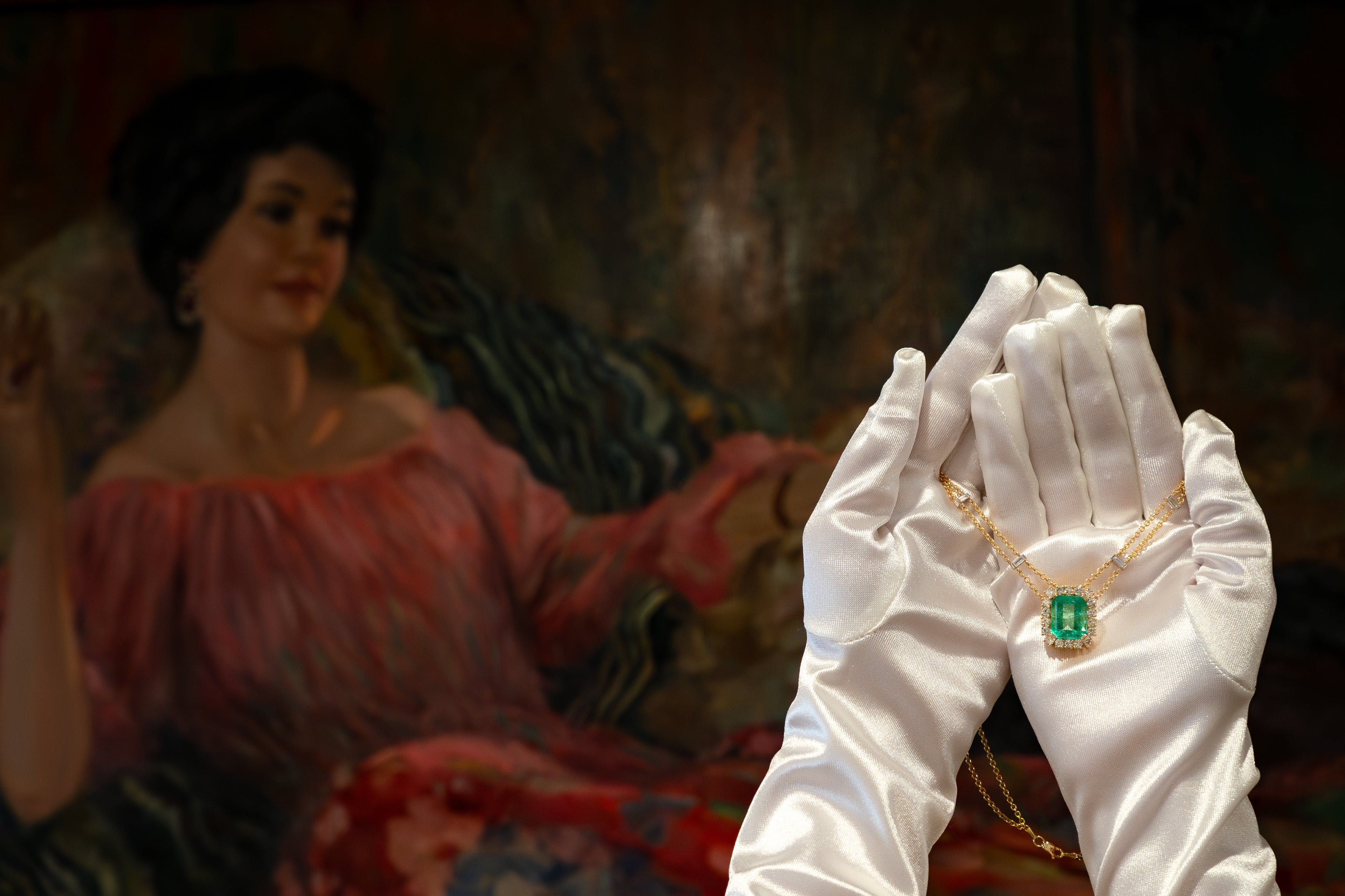 White-gloved hands holding a gold necklace with a green gemstone in front of a classical painting.